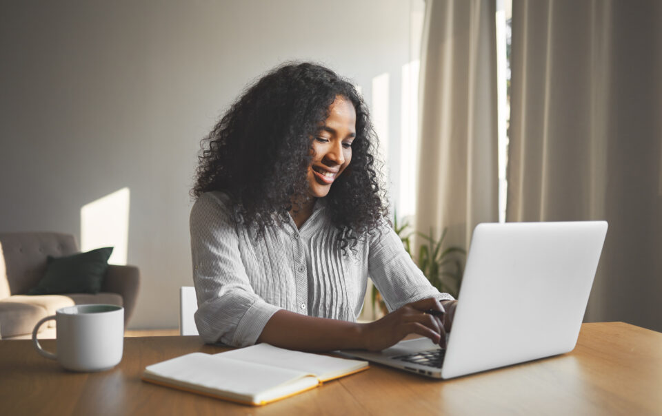 gorgeous positive young dark skinned female blogger keyboarding generic laptop smiling being inspired while creating new content her travel blog sitting desk with diary mug