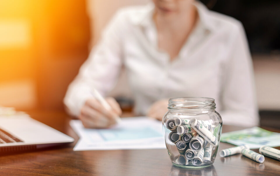 a jar with rolled banknotes on the table laptop papers woman on the background