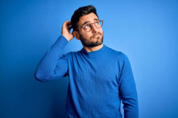 young handsome man with beard wearing casual sweater glasses blue background confuse wondering about question uncertain with doubt thinking with hand head pensive concept